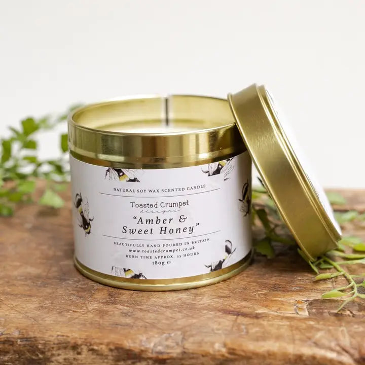 Candle tin labeled 'Amber & Sweet Honey' on a wooden surface with a blurred green plant background