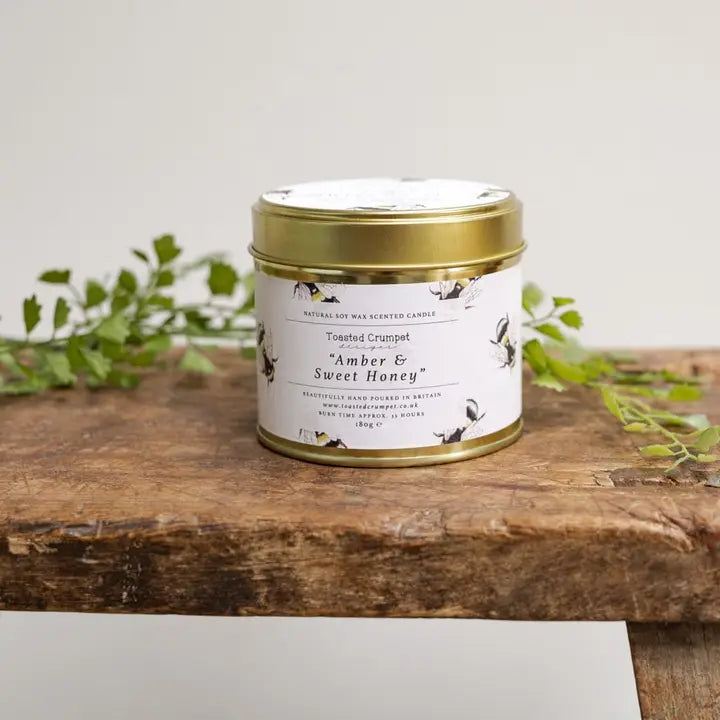 Candle in a tin labeled 'Amber & Sweet Honey' on a wooden surface with greenery in the background.