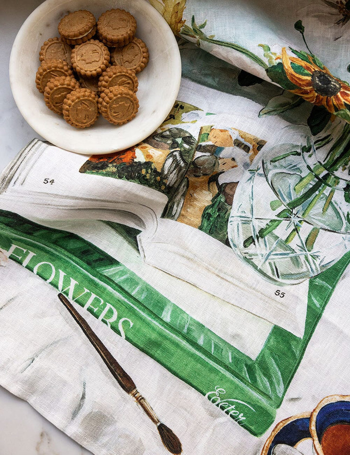 Bowl of cookies on a floral-patterned cloth with an open book and paintbrush.