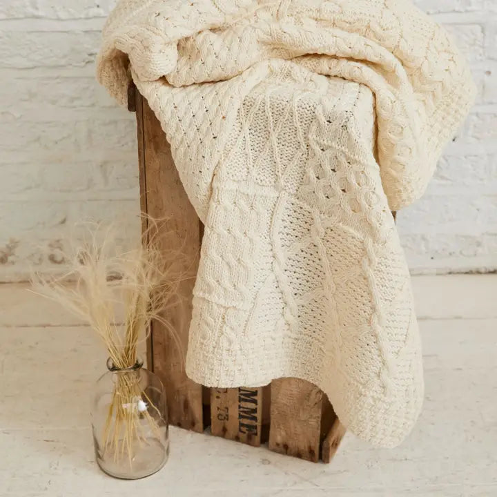 Knitted beige blanket draped over a wooden crate with a vase of dried plants on a light wooden floor.