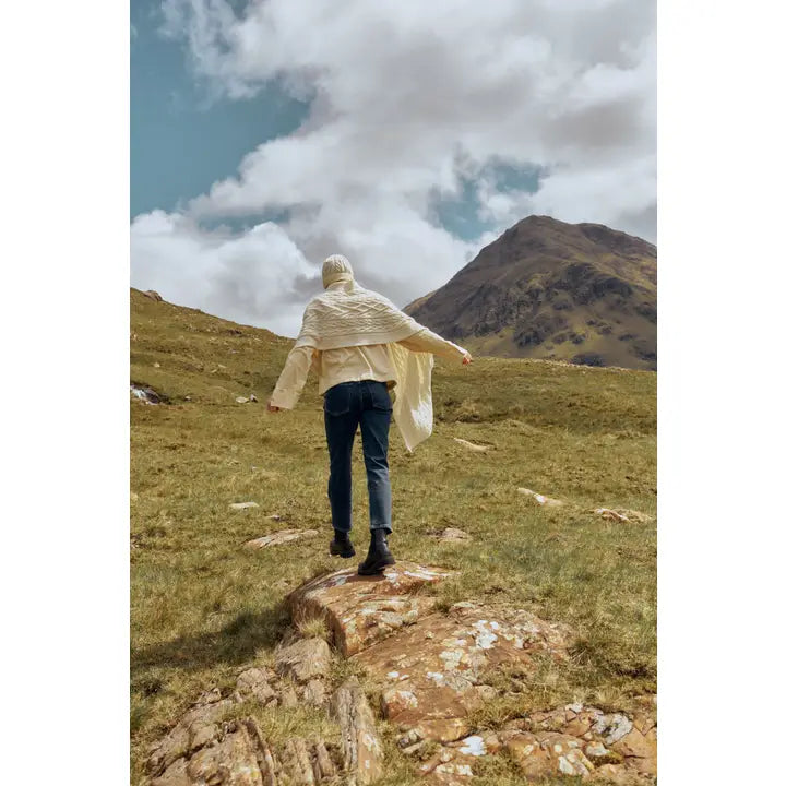 Person walking on a rocky path with mountains in the background wearing jeans a cream scarf