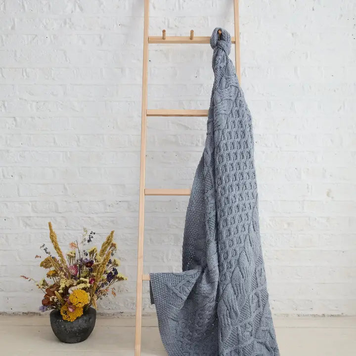 Gray Blue textured blanket draped over a wooden ladder against a white brick wall with a small potted plant.