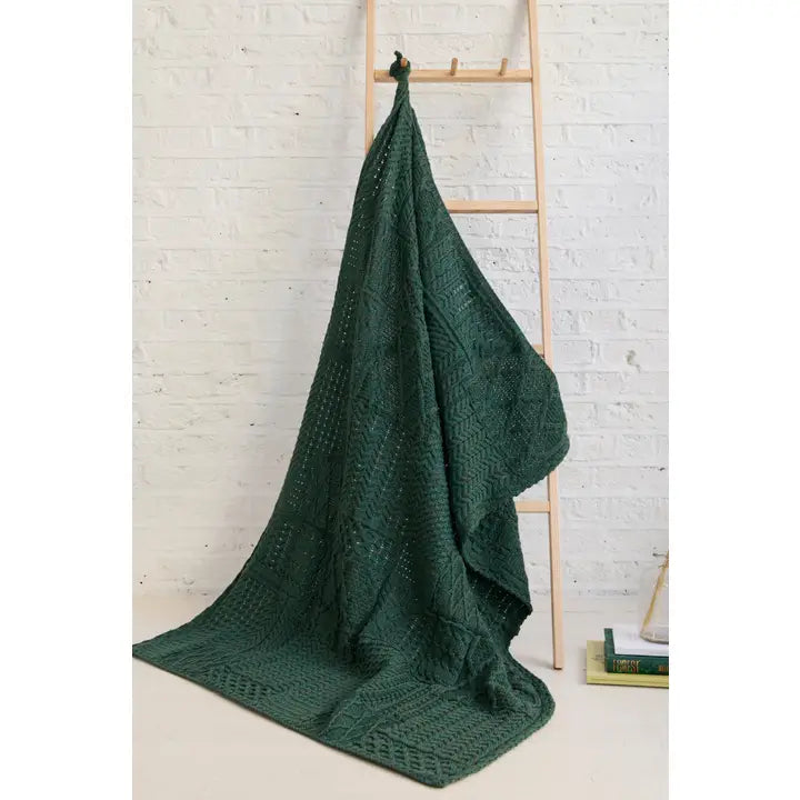 Green knitted blanket draped over a wooden ladder against a white brick wall.