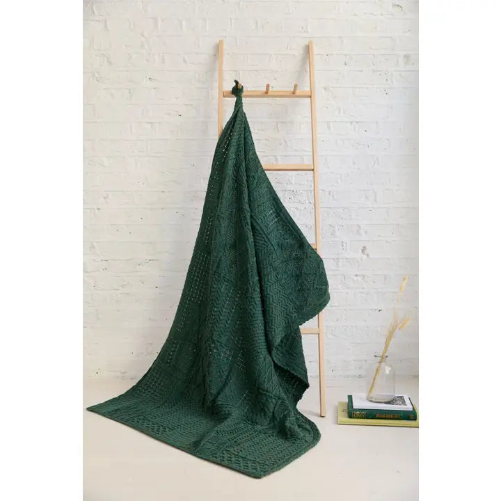 Green knitted blanket draped over a wooden ladder against a white brick wall.