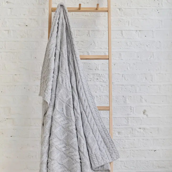 Gray textured blanket draped over a wooden ladder against a white brick wall.
