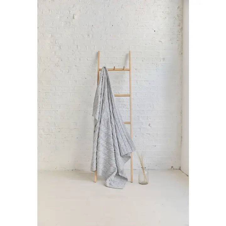 Gray textured blanket draped over a wooden ladder against a white brick wall.