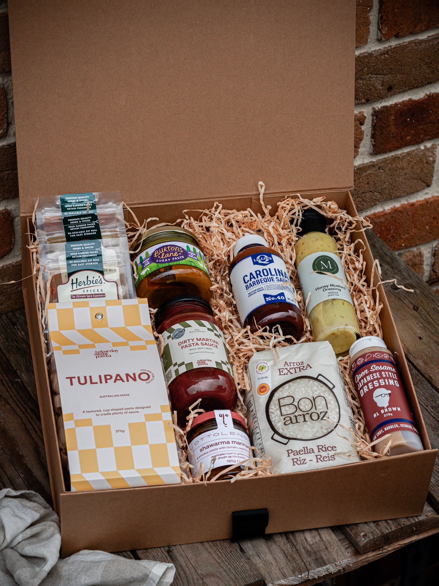 Gift box with various food products including sauces and a bottle of olive oil on a wooden surface.