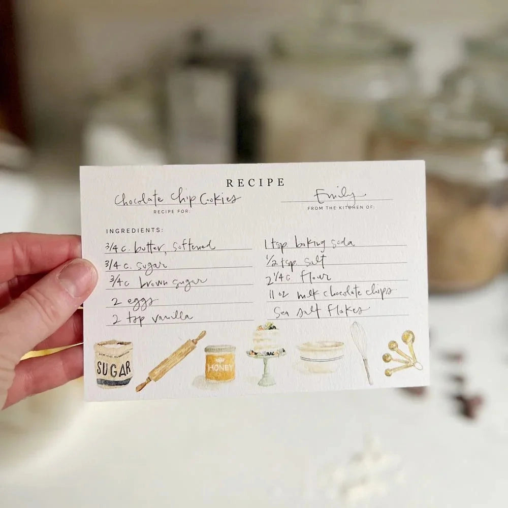 Hand holding a recipe card for chocolate chip cookies with kitchen utensils on a blurred background.