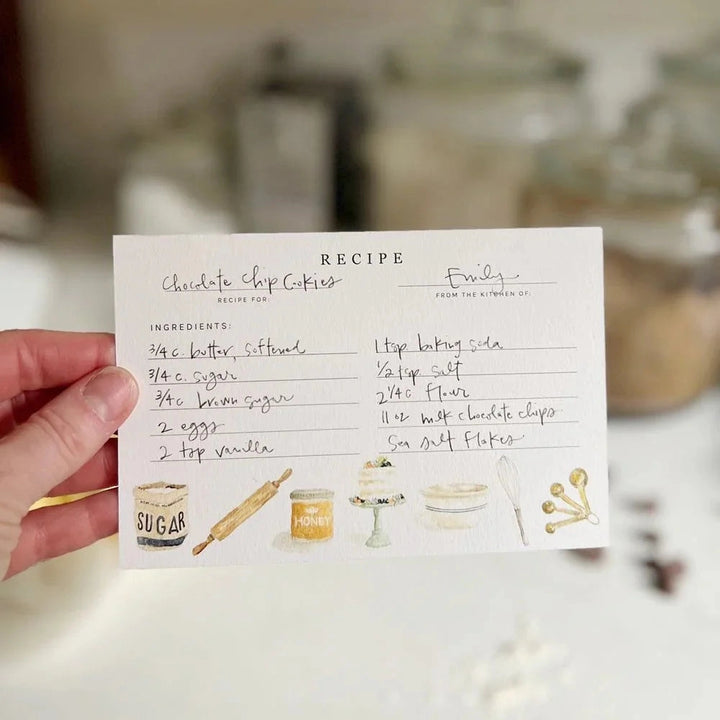 Hand holding a recipe card for chocolate chip cookies with kitchen utensils on a blurred background.