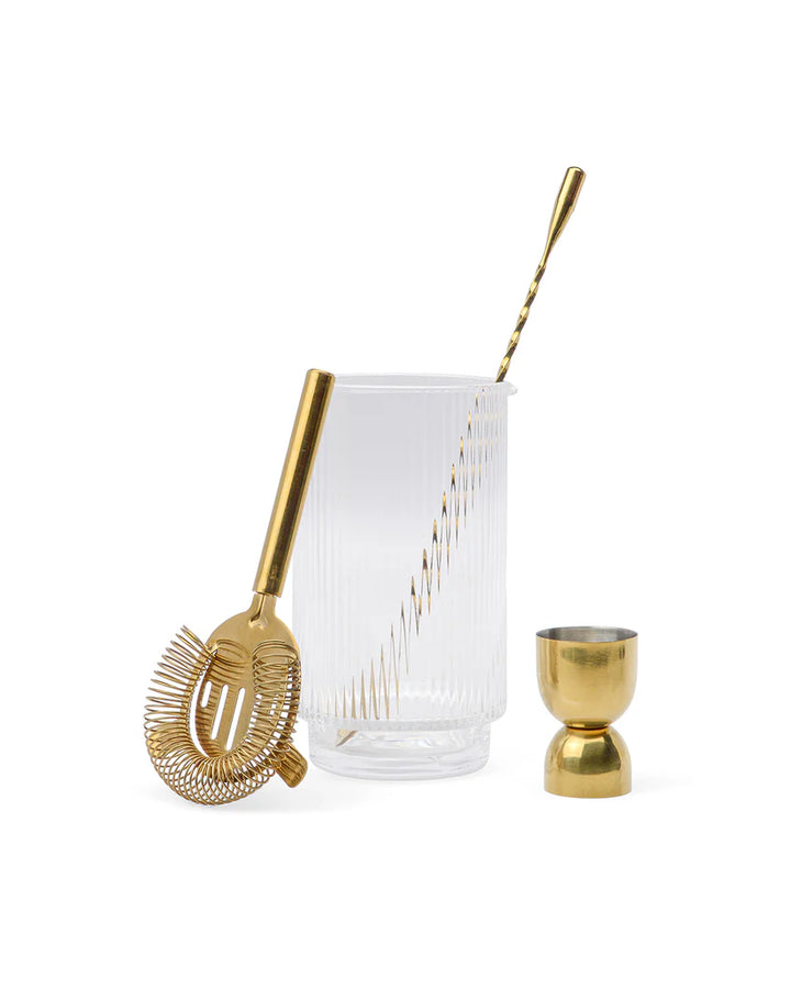 A bar tool set including a gold-plated stainless steel cocktail shaker, a fluted beaker in clear soda lime glass, and a gold-plated jigger, all arranged neatly on a white background.