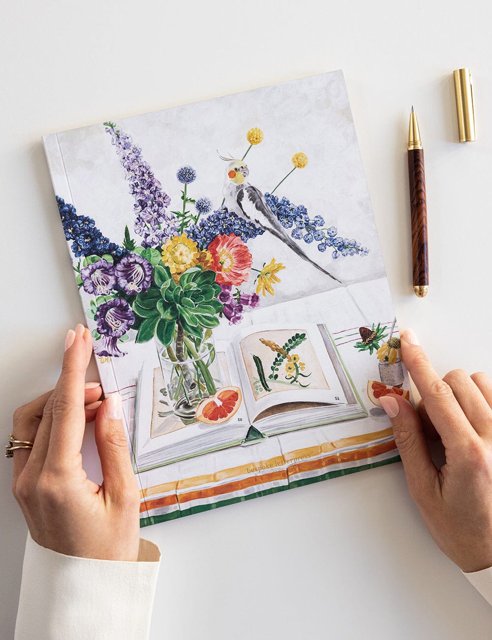 Notebook with floral illustrations held by hands on a white surface with a pen.