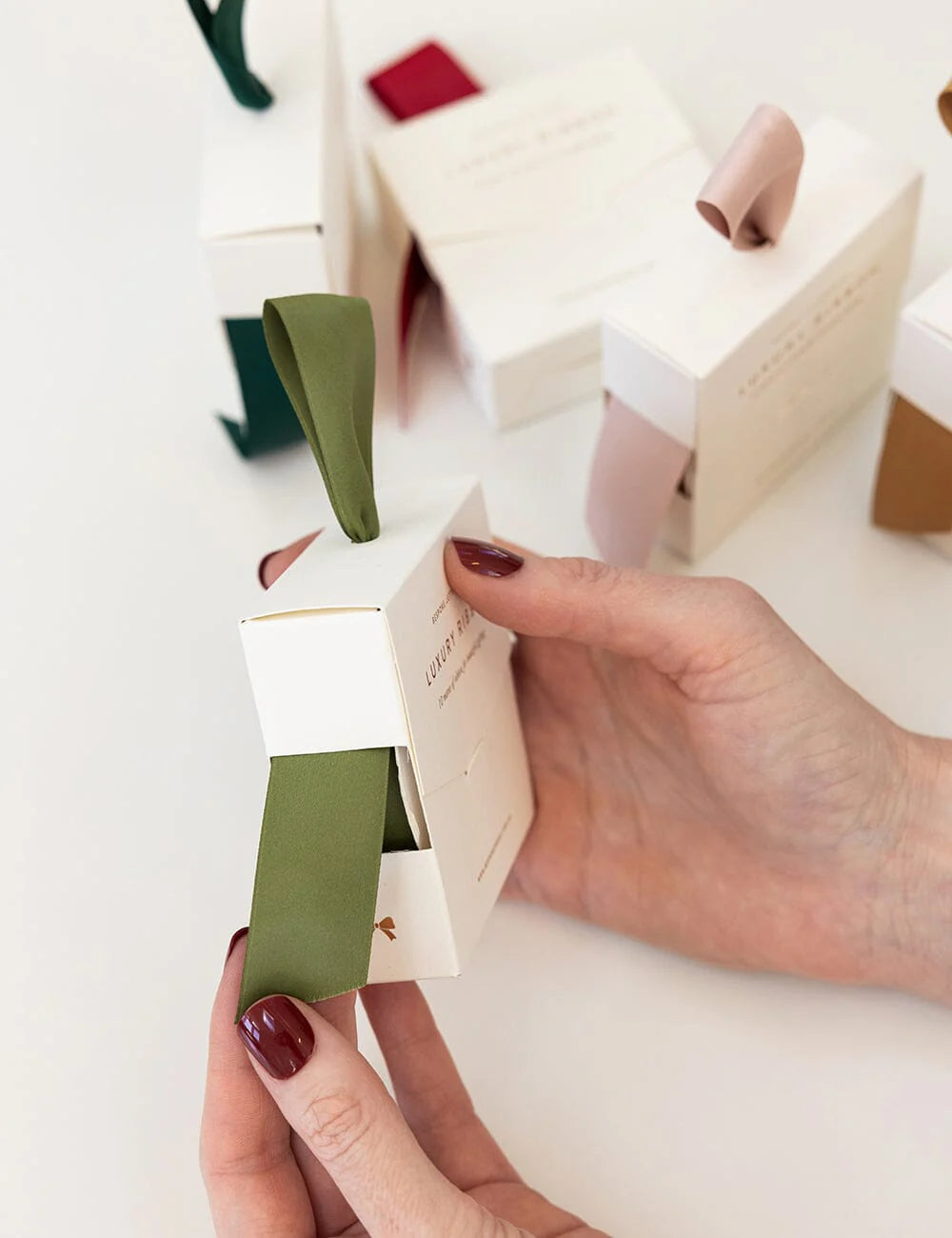 Person holding a small white box with olive satin ribbon inside on a light background