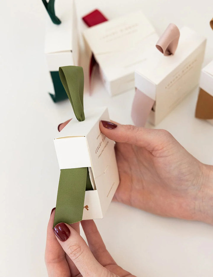 Person holding a small white box with olive satin ribbon inside on a light background
