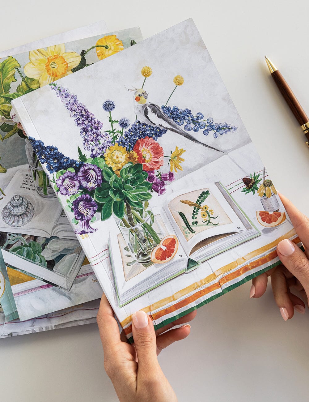 Colorful floral illustrations on a book held by hands with a pen nearby.