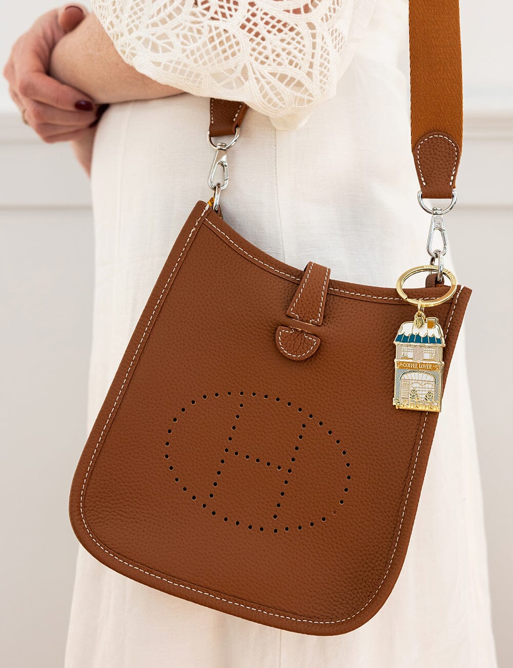 Brown leather handbag with a distinctive logo and a gold charm, held by a person wearing a white lace top.
