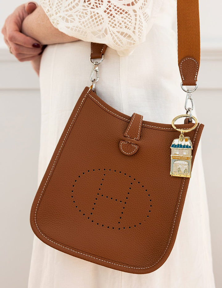 Brown leather handbag with a distinctive logo and a gold charm, held by a person wearing a white lace top.