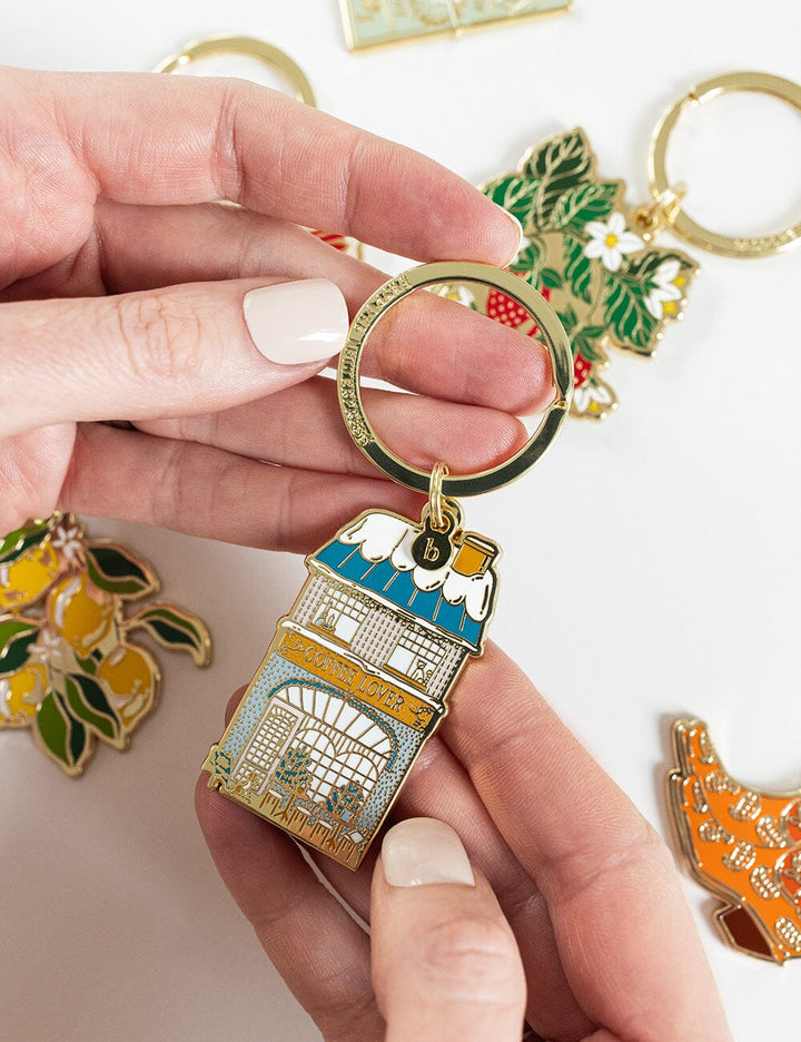 Hand holding a decorative keychain with a building design, surrounded by other similar keychains on a light background.