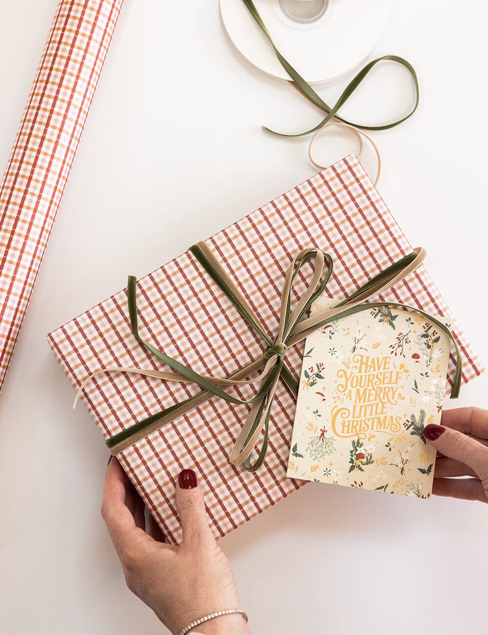 Gift box with red and white checkered paper, green and gold ribbon, and a Christmas-themed card on a light background.