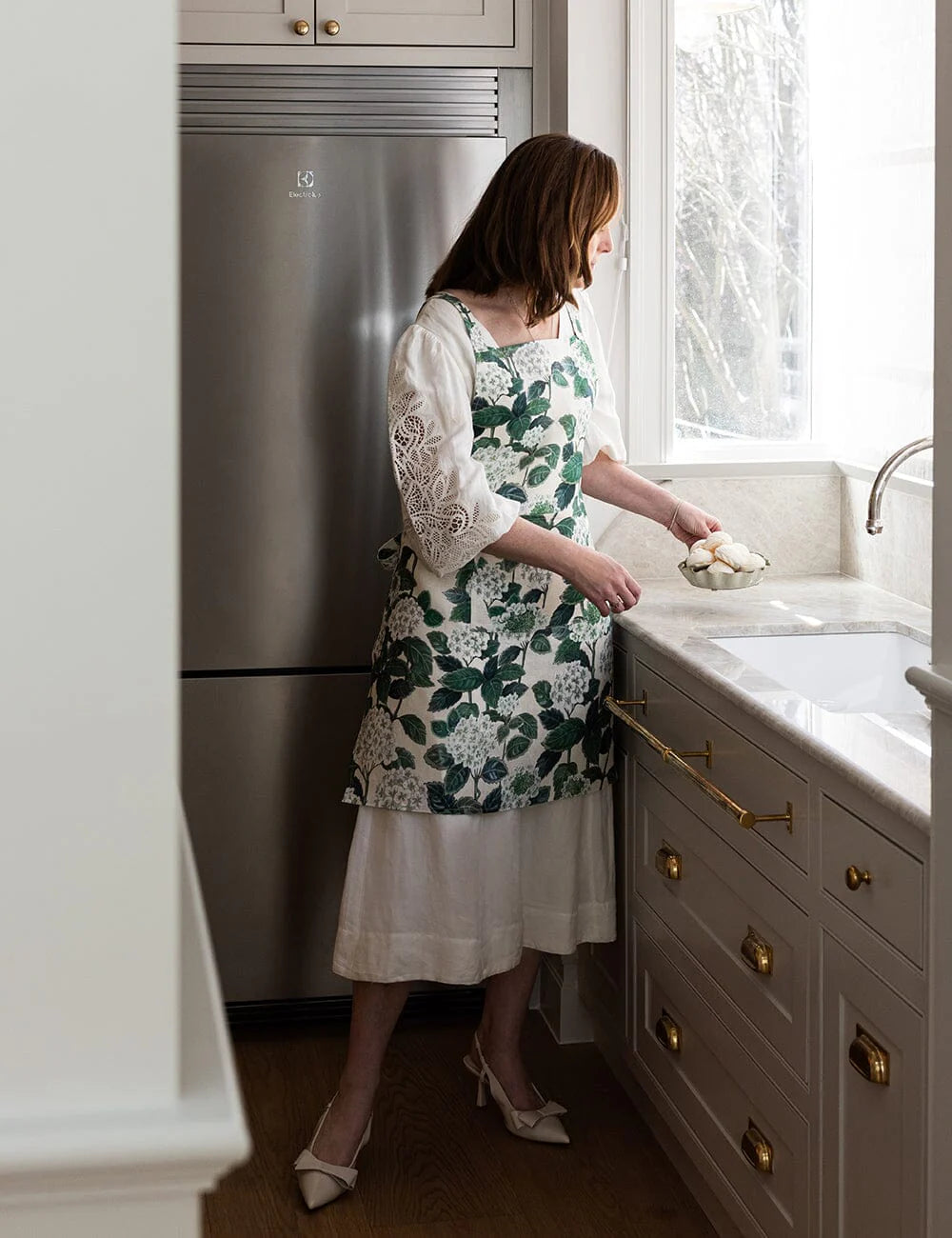 Woman in a floral apron standing in a kitchen next to a sink.