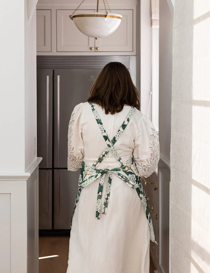 Woman in a white dress with a floral apron standing in a kitchen.