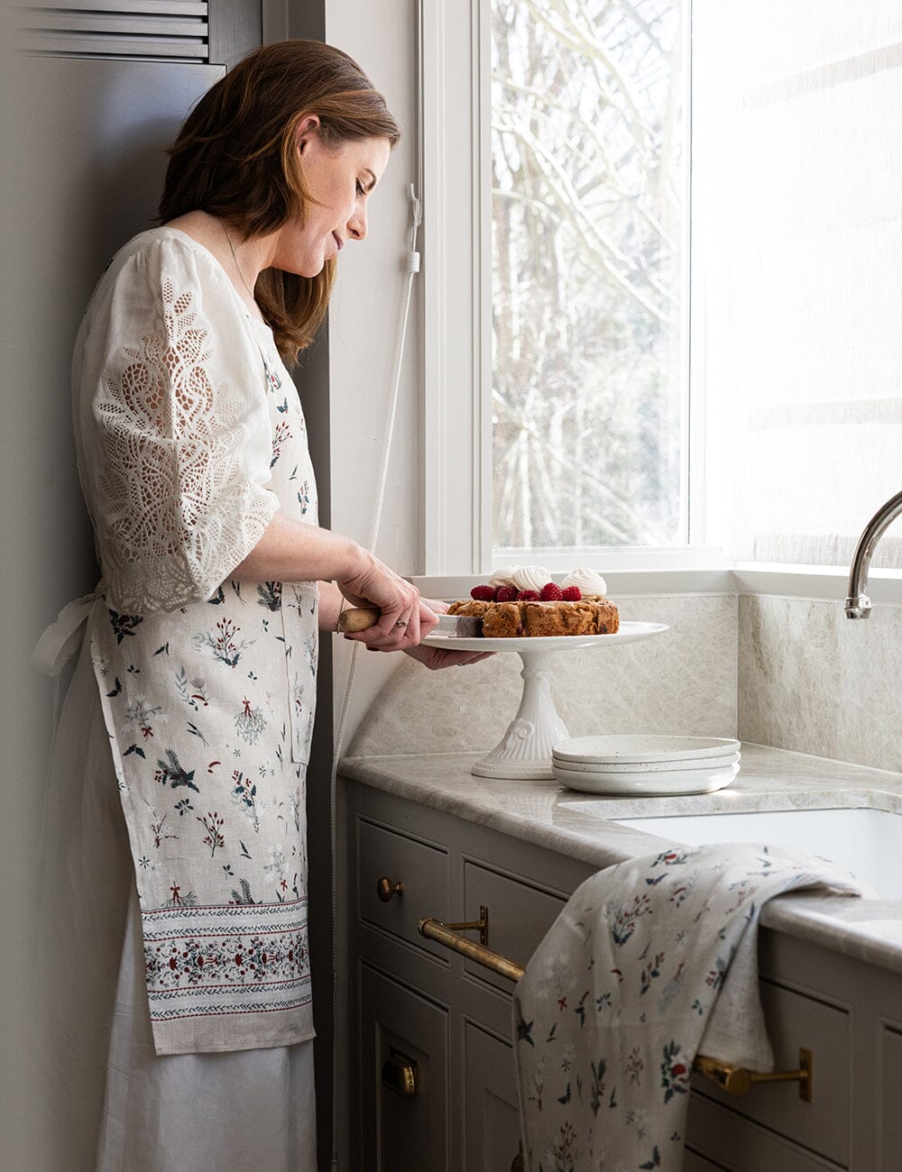 Woman in a kitchen holding a cake, wearing a floral apron.