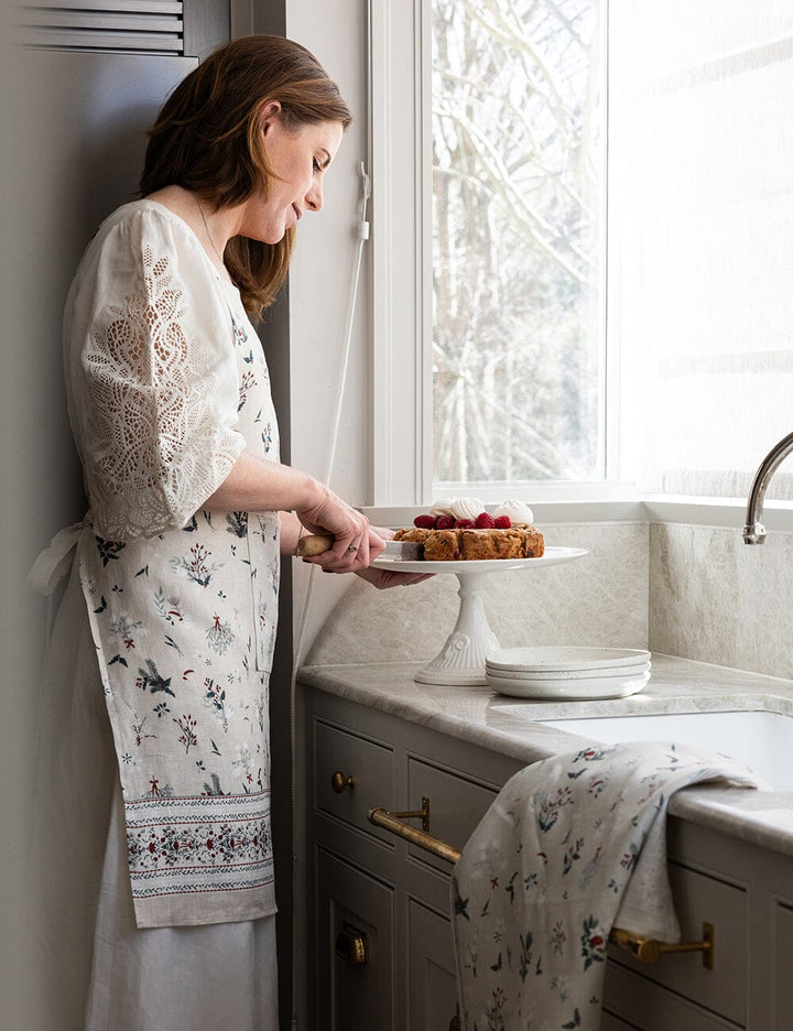 Woman in a kitchen holding a cake, wearing a floral apron.