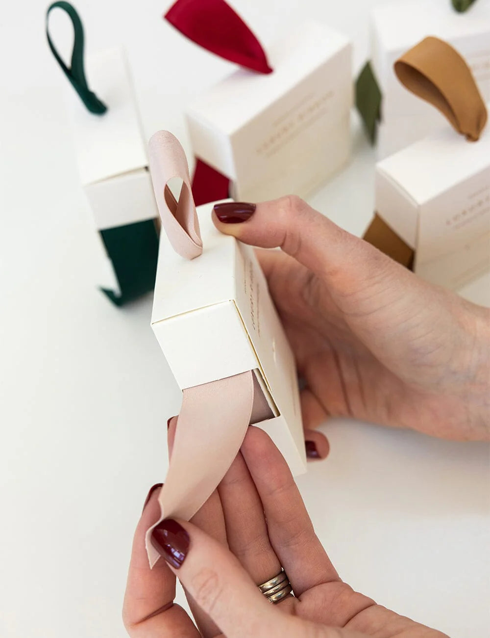 Person holding a small white box with a nude bow against a blurred background of gift boxes.