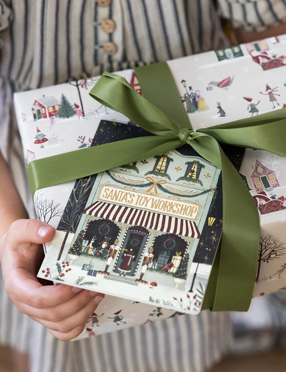 Gift box with 'Santa's Toy Workshop' design and green ribbon held by a person wearing a striped shirt.