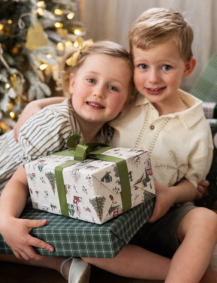 Two children holding a stack of Christmas presents in front of a decorated tree.
