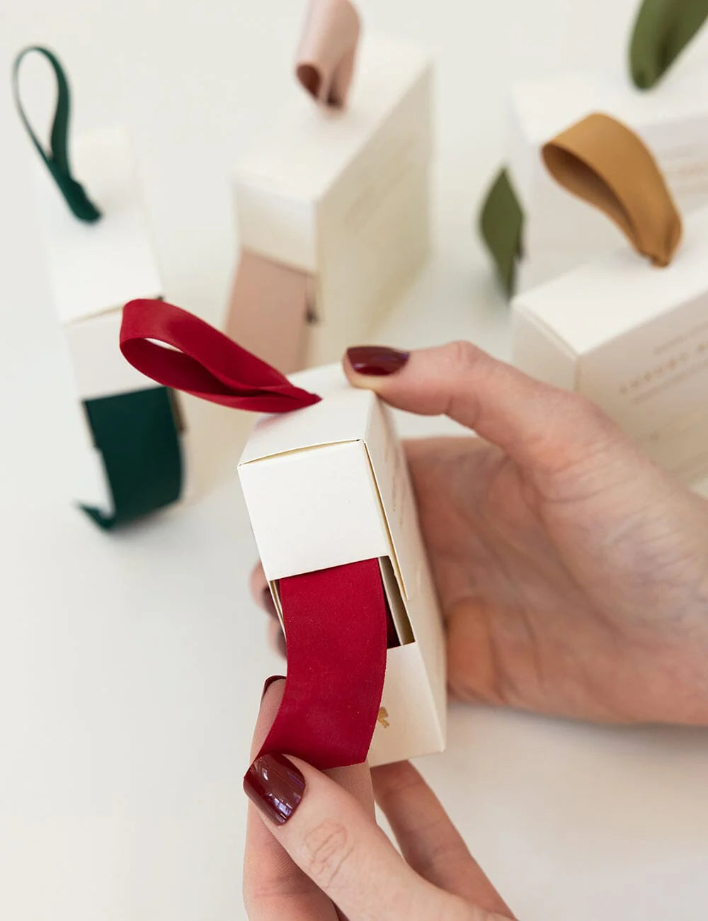 Hand holding red ribbon in white packaging with blurred gift boxes in the background