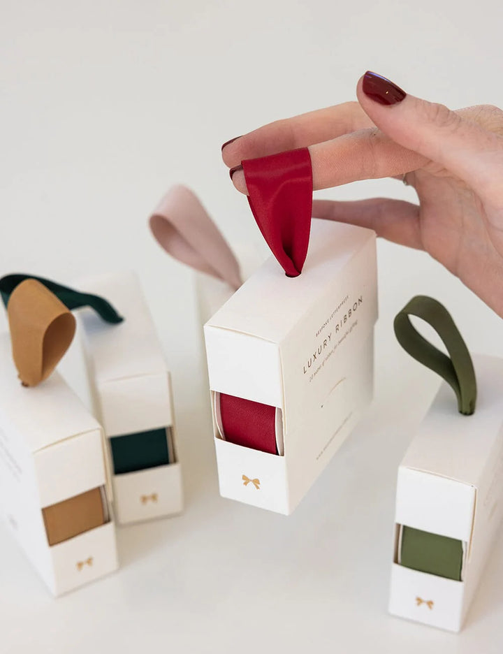 Hand holding a red ribbon over a white box with a brand name, surrounded by other similar boxes.