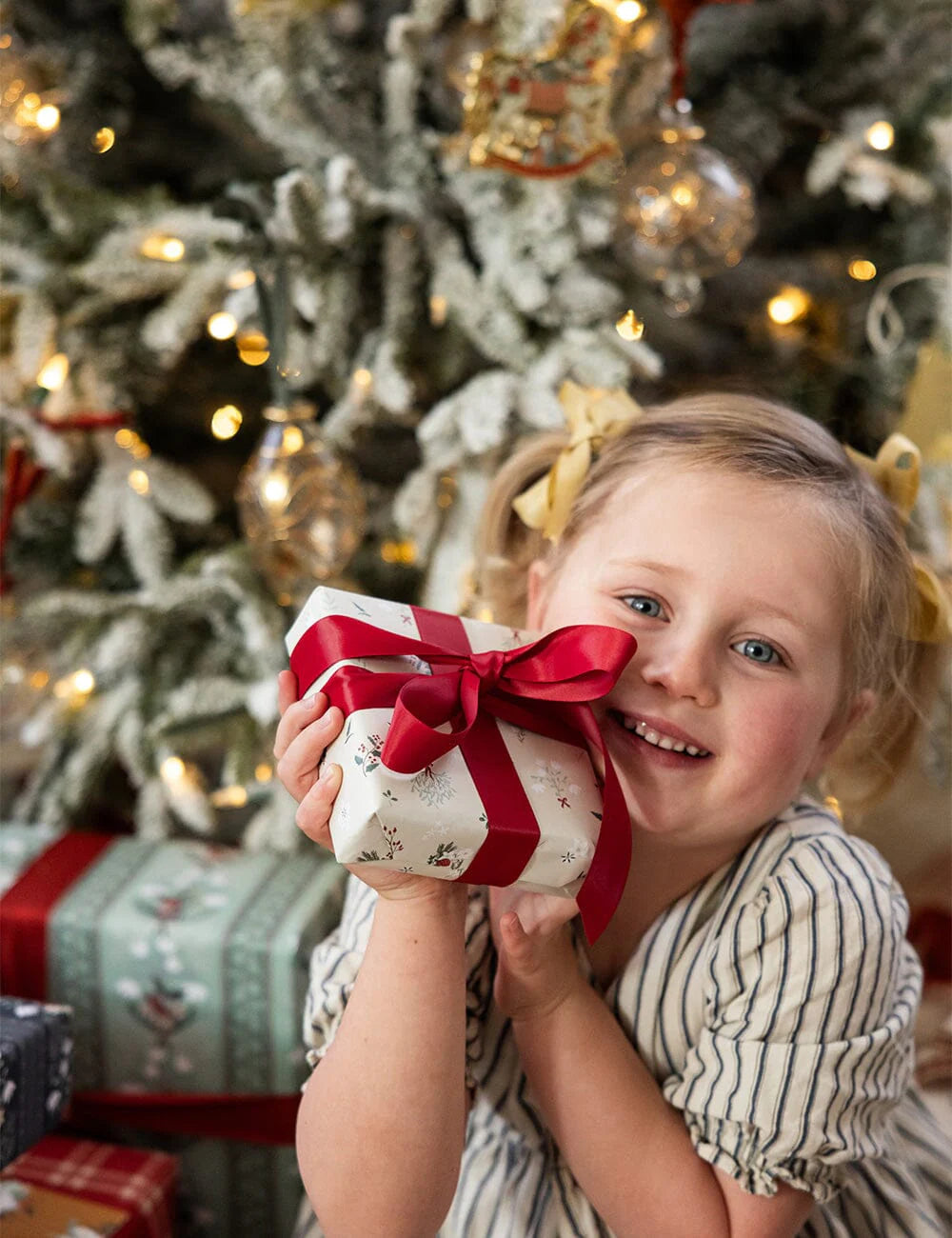Child holding a gift with a red ribbon in front of a decorated Christmas tree.