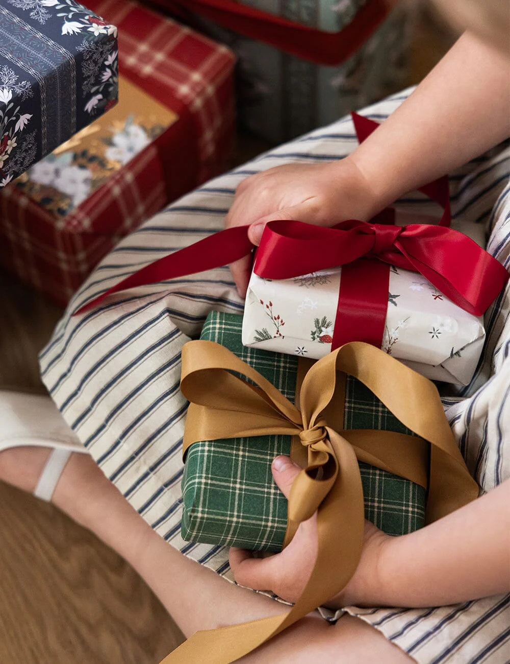 Person holding a stack of wrapped gifts with ribbons on a striped surface