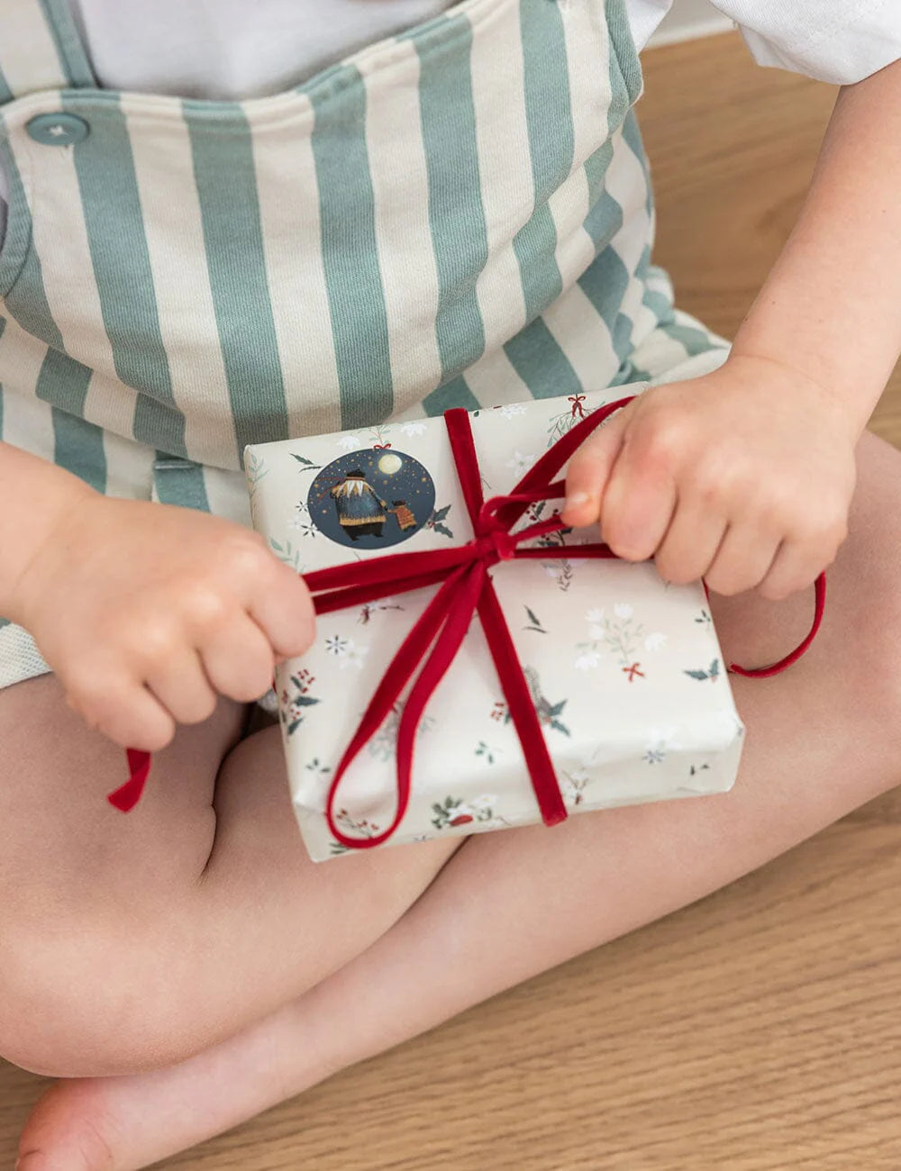 Child's hands holding a wrapped gift with a red ribbon on a wooden floor.