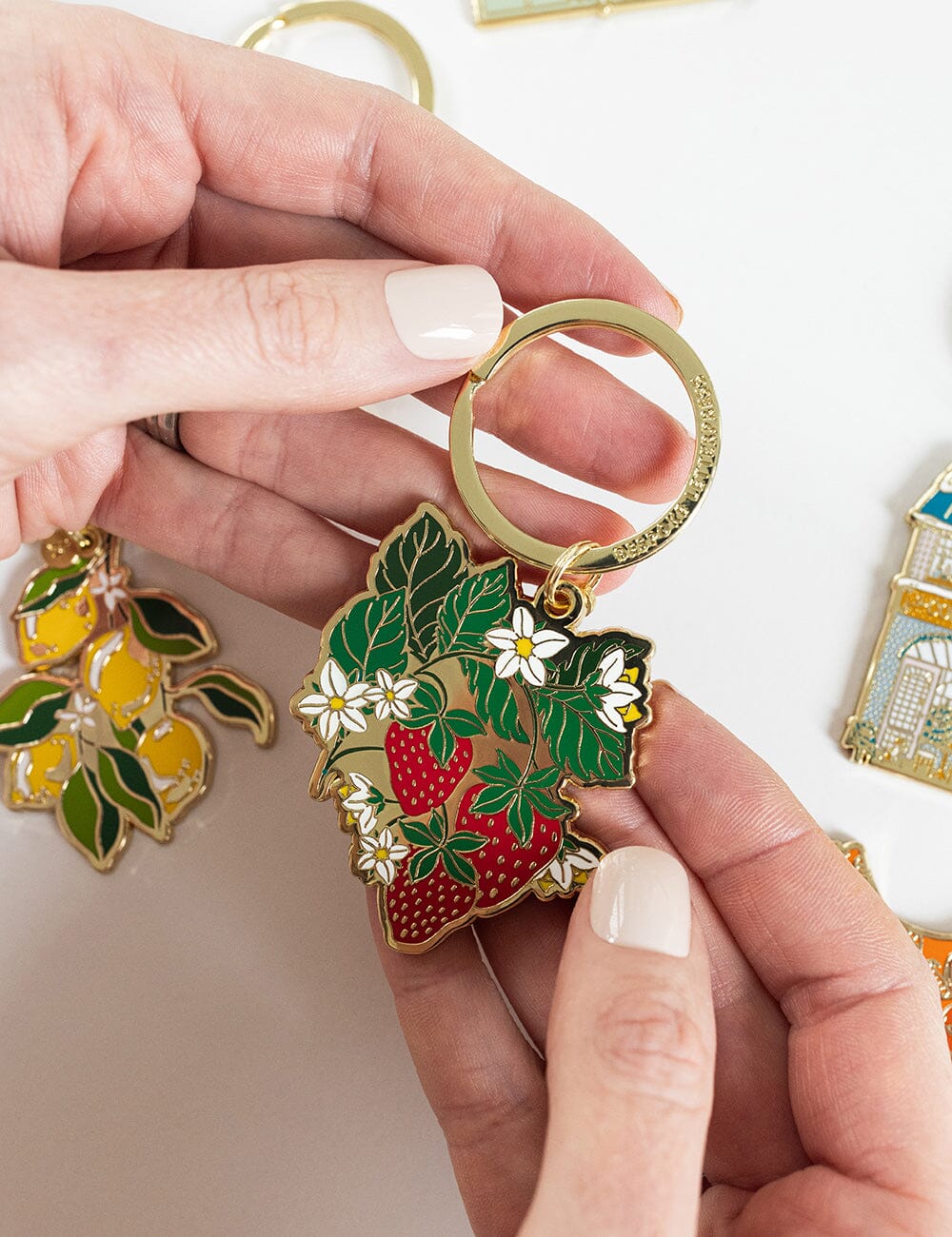 Hand holding a keychain with strawberry and leaf design, surrounded by other decorative items.