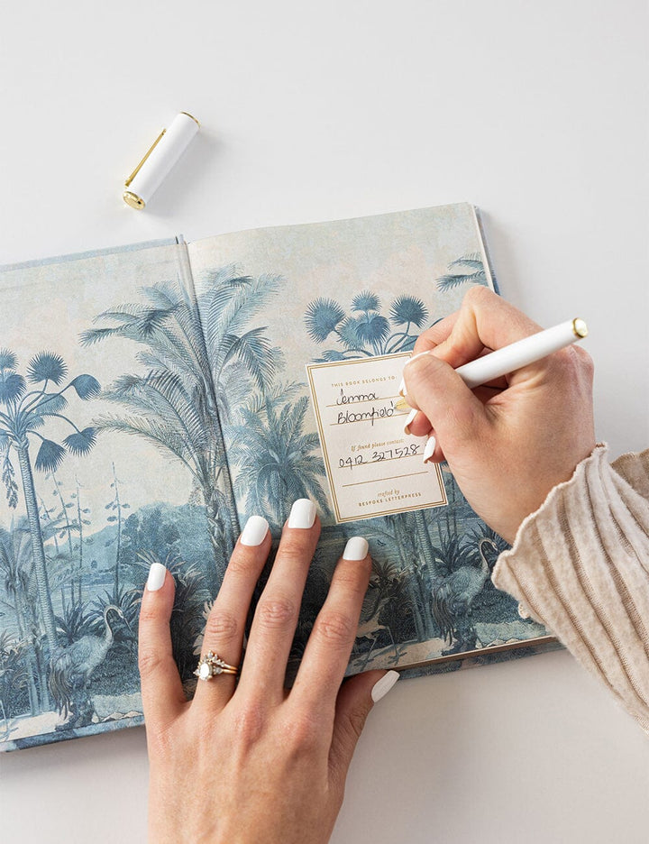 Person writing in a notebook with a scenic cover on a light surface