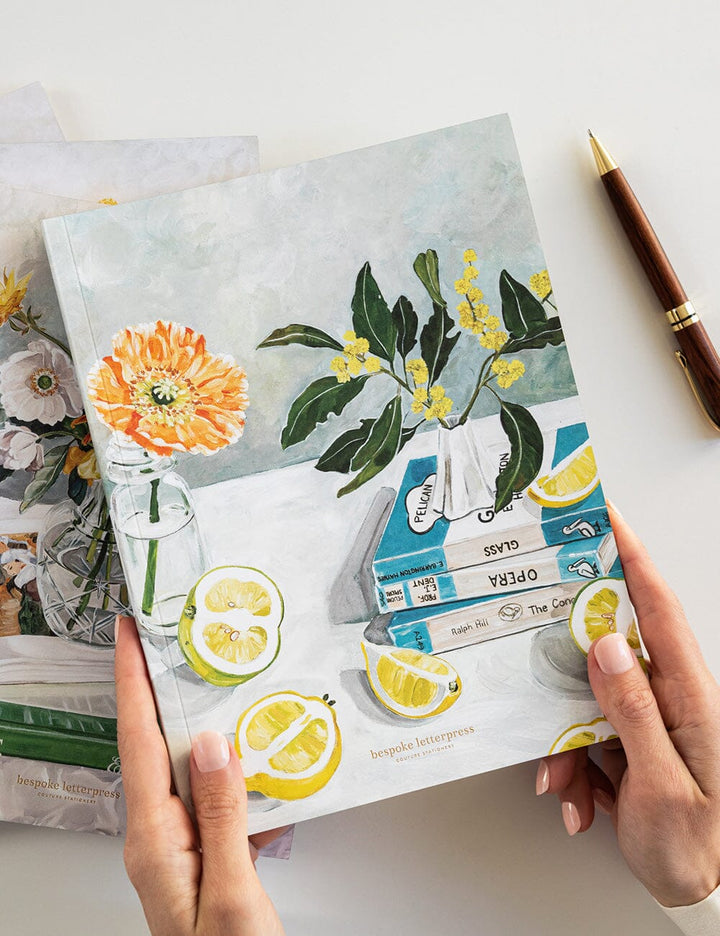 Notebook with floral and lemon illustrations held by hands on a light surface.