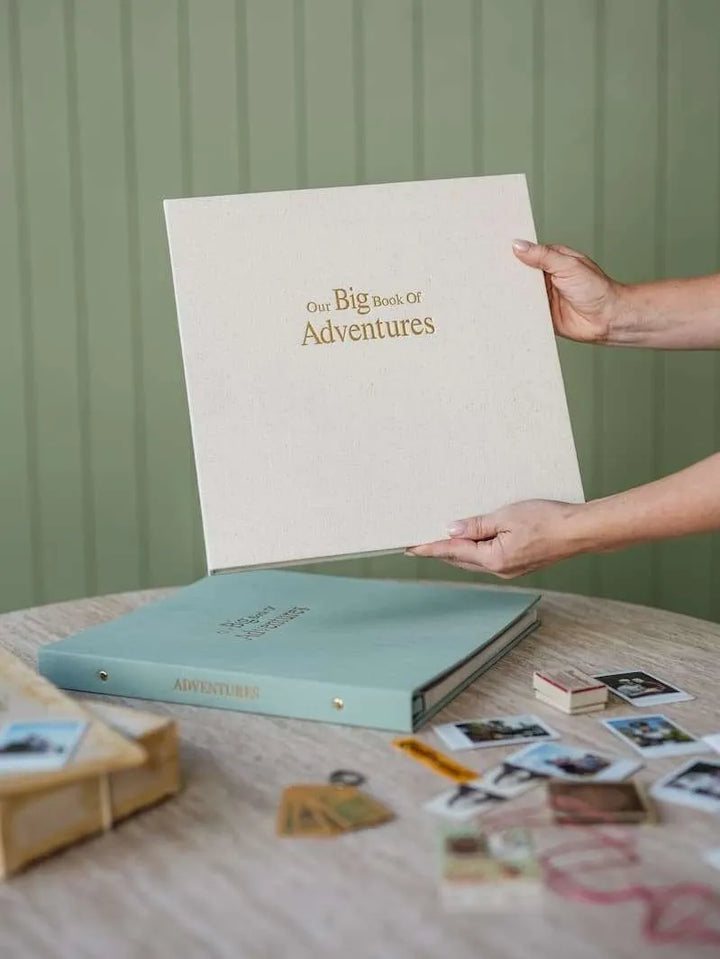 Person holding a large book titled 'Our Big Book of Adventures' with a smaller blue book and photos on a table.