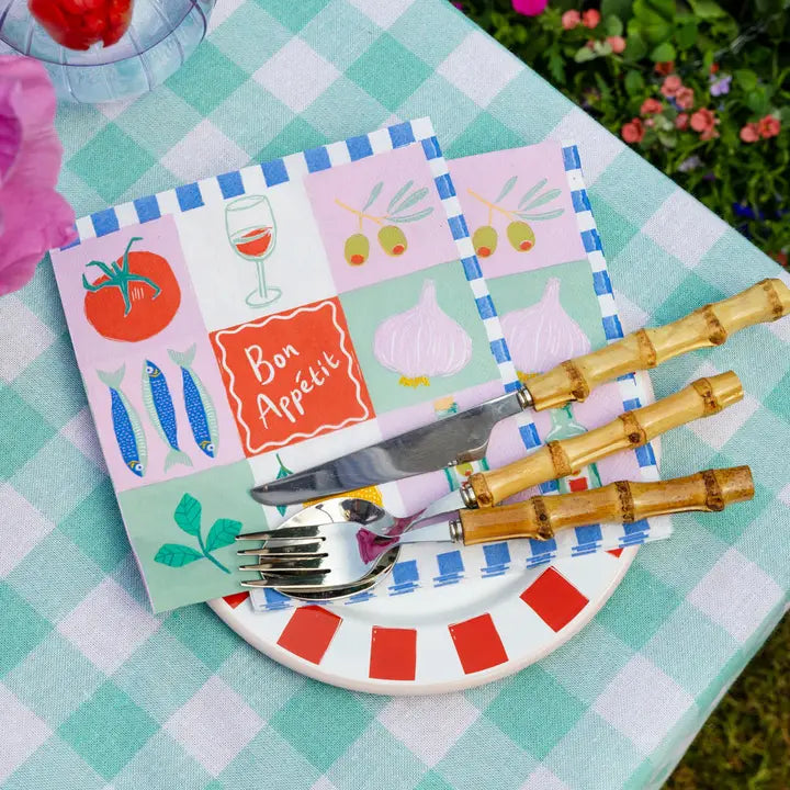 Set of cutlery with bamboo handles on a colorful plate with a 'Bon Appetit' card on a checkered tablecloth.