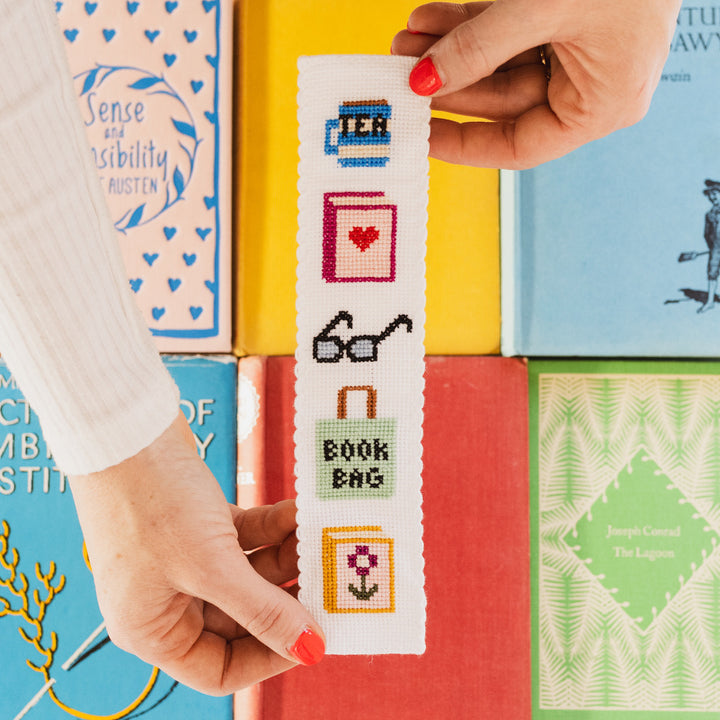 Hand holding a cross-stitched bookmark with book-themed designs against a background of colorful books.