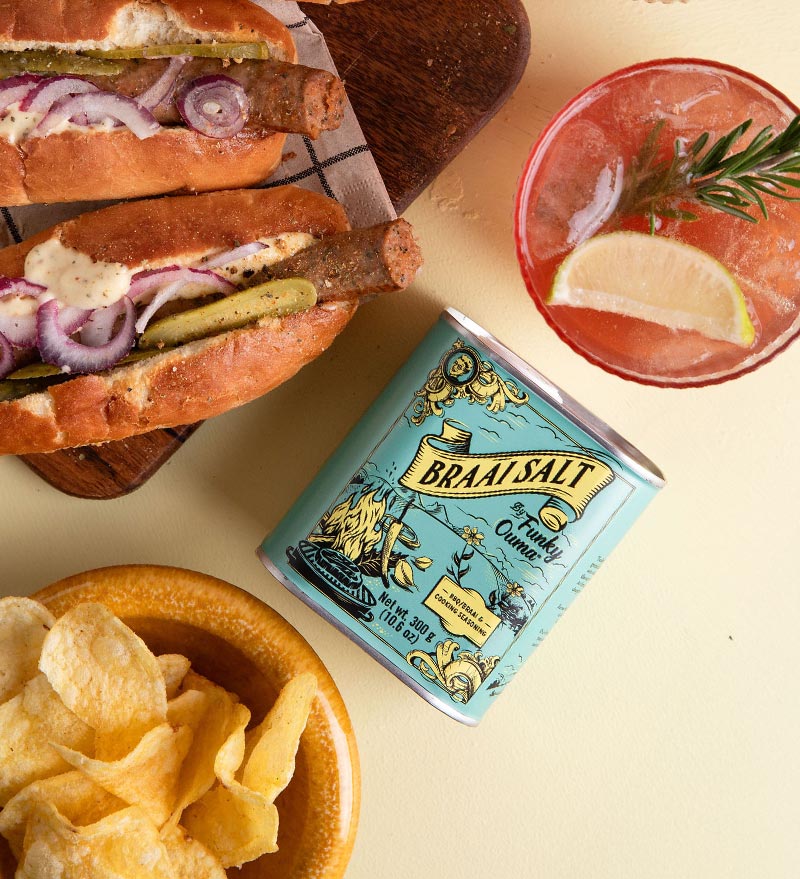 Braai Salt can with two hot dogs, chips, and a cocktail on a yellow background