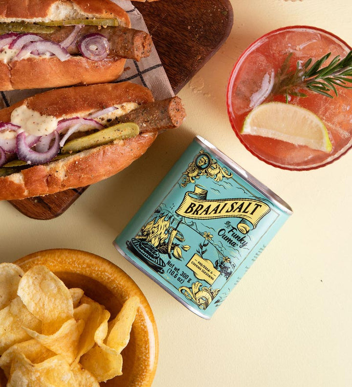 Braai Salt can with two hot dogs, chips, and a cocktail on a yellow background