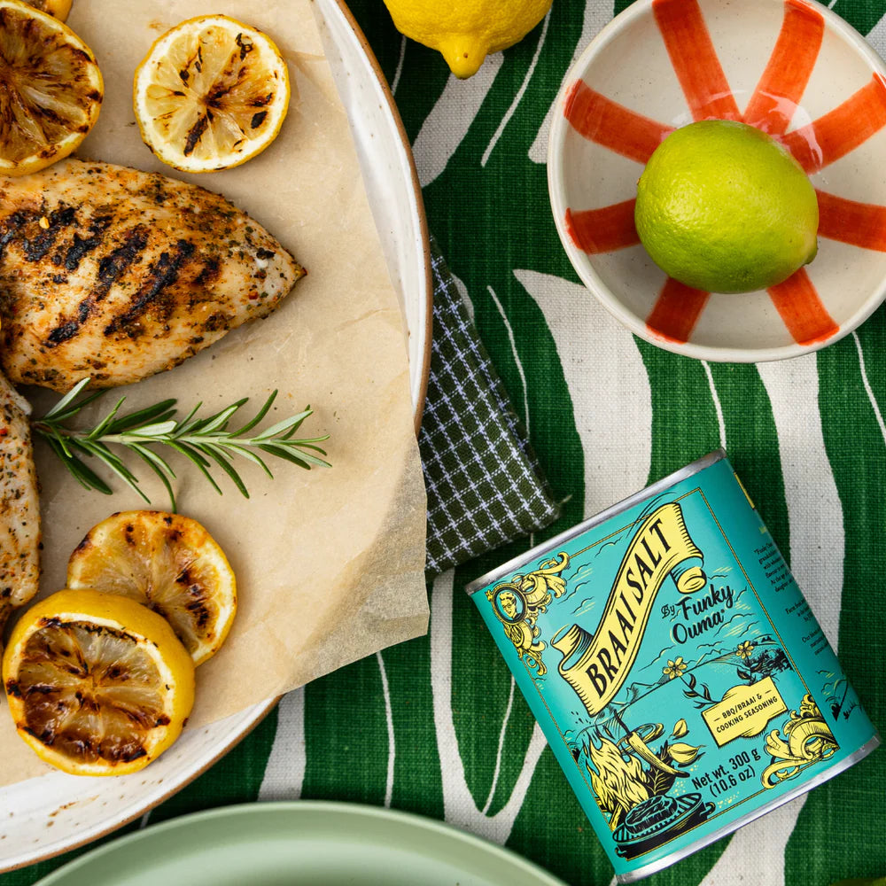 Grilled chicken with lemons and a tin of Braai Salt on a green tablecloth.