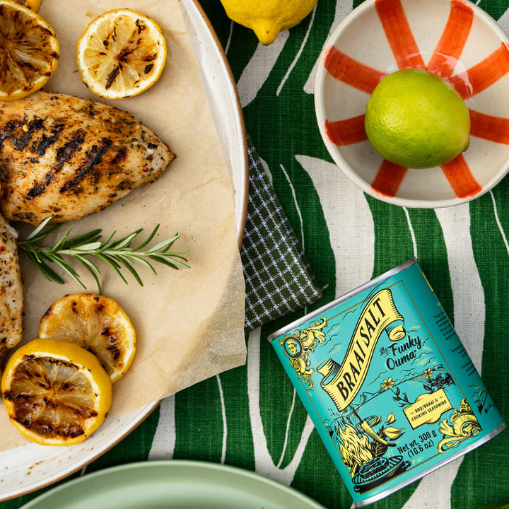Grilled chicken with lemons and a tin of Braai Salt on a green tablecloth.