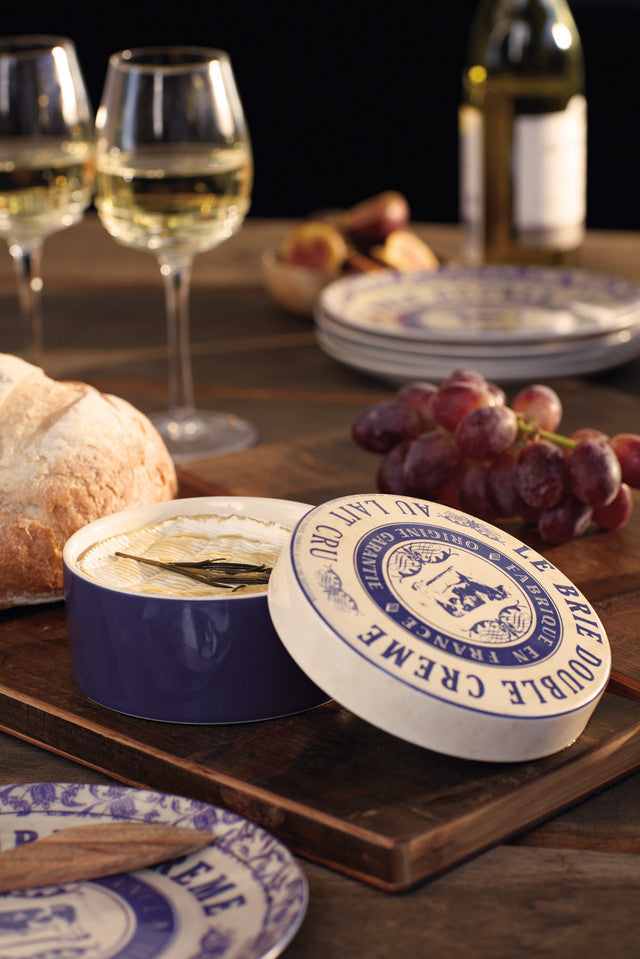 Ceramic cheese baker with blue and white labels on a wooden board with wine glasses and grapes.