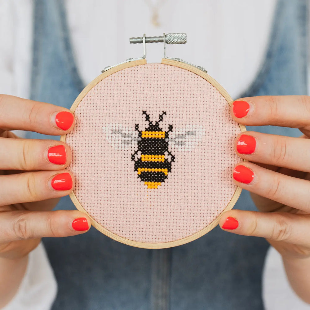 Cross-stitched bee design on fabric held by hands with red nail polish.