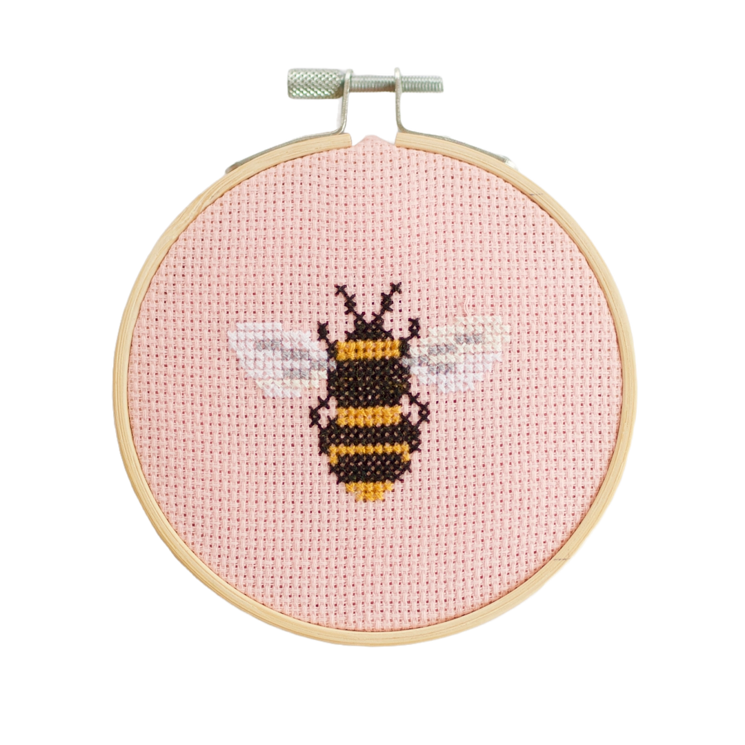 Cross-stitched bee design on a pink background in a wooden hoop.
