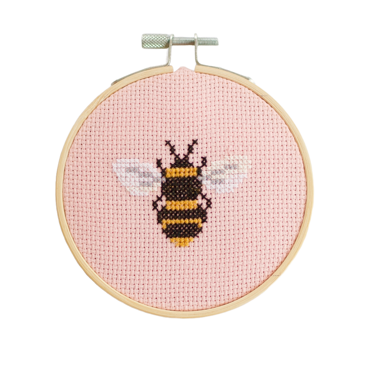 Cross-stitched bee design on a pink background in a wooden hoop.