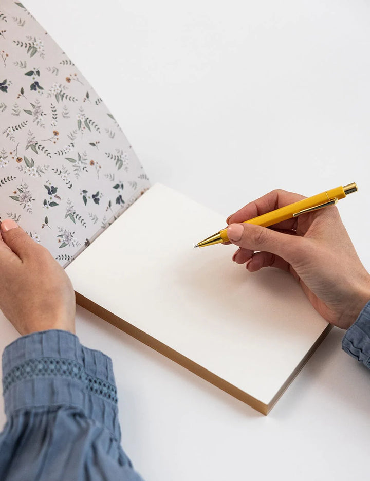 Person writing in a notebook with a yellow pen on a white surface