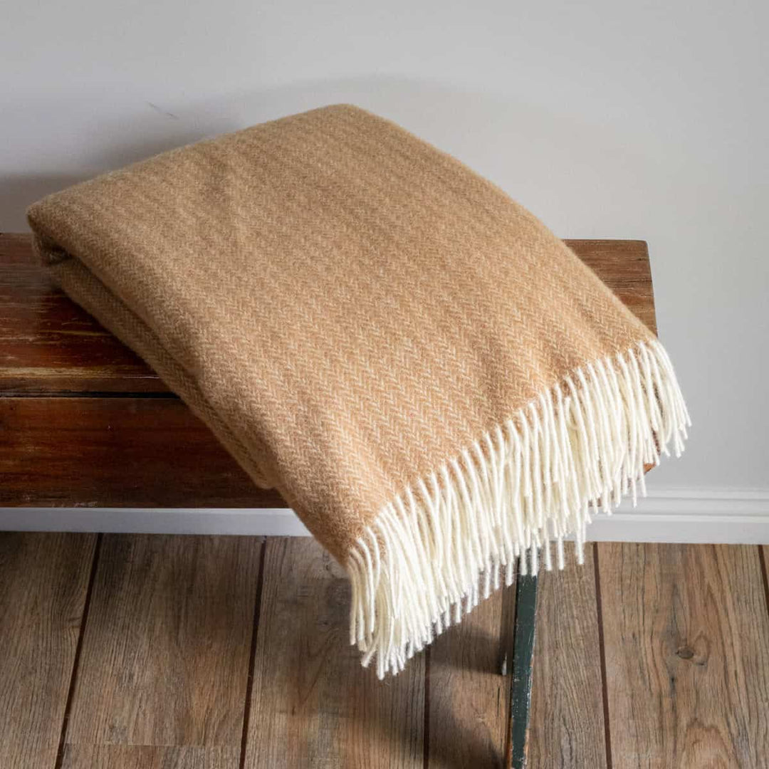 Beige blanket with white fringe draped over a wooden bench against a neutral wall.